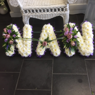 Nan Tribute - Beautiful white Chrysanthemums with a flourish of Lillac/pink Roses and purple Eustomia, edged with red ribbon pleats, form the name NAN.

Stand included.

Please note this tribute is availble in several colour choices, please advise when placing your order.

Purple, Lilac, Purple, Yellow, Cream. Blue, Orange, Red

Please note this item is only suitable for delivery to a funeral. Please make a selection from an alternative range, if you would like to express your sympathy to the bereaved. Product photography is indicative of the highest price point. Please note that seasonal availability of individual stems may vary from those in the picture. Our professional florist may substitute flowers for a suitable alternative, similar in style, colour, quality and value. Where flower designs include a sundry item such as a vase or basket it may not always be possible for our florists to include the exact item pictured. Every effort will be taken to ensure that the item is substituted for a similar design.Price above includes delivery charge but excludes relay fee.